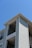 The exterior of a light-colored, multi-story residential building with siding and balconies features prominently. The architecture includes clean lines and a modern design, with visible railings on the upper floors. The building is set against a clear blue sky, suggesting a sunny day.