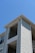 The exterior of a light-colored, multi-story residential building with siding and balconies features prominently. The architecture includes clean lines and a modern design, with visible railings on the upper floors. The building is set against a clear blue sky, suggesting a sunny day.
