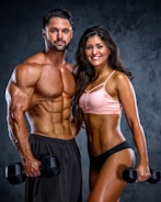 A muscular man and woman are posing confidently with dumbbells in their hands. The man is shirtless wearing black shorts, while the woman is wearing a pink sports bra and black shorts. Both display impressive physiques against a dark textured background.
