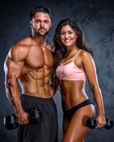 A muscular man and woman are posing confidently with dumbbells in their hands. The man is shirtless wearing black shorts, while the woman is wearing a pink sports bra and black shorts. Both display impressive physiques against a dark textured background.