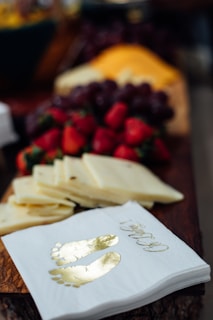 A close-up of a wooden board featuring sliced cheese, fresh strawberries, grapes, and decorative napkins with gold footprints printed on them.