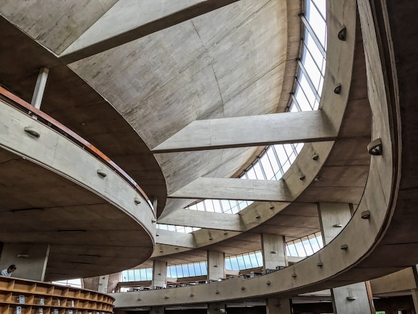 A modern architectural interior with large, sweeping concrete curves and expansive high windows allowing natural light to flood the space. The design features multiple levels and a spacious open layout, creating an airy and dynamic atmosphere.