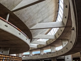 A modern architectural interior with large, sweeping concrete curves and expansive high windows allowing natural light to flood the space. The design features multiple levels and a spacious open layout, creating an airy and dynamic atmosphere.