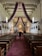 A person walks down the aisle of a church, which features high wooden ceilings with purple and pink drapes hanging. The church has two rows of wooden pews, intricate wall decorations, and a richly adorned altar area with religious artifacts.