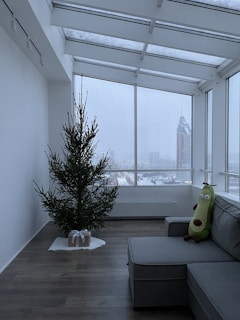 A minimalist living room with a large window offering a view of a snowy cityscape. An undecorated Christmas tree stands on the wooden floor with a single gift wrapped in brown paper beneath it. A grey sectional sofa is adorned with a plush toy resembling a cartoonish character or vegetable.