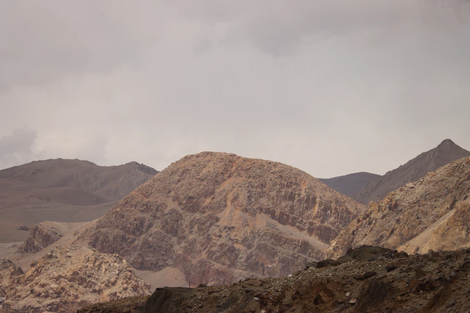 The image depicts a mountainous landscape with a large, jagged rock formation on the left side and smaller hills on the right side. The sky above is overcast, casting a soft light over the scene. The colors are predominantly brown and gray, reflecting the rocky terrain of the mountains and the cloudy sky.