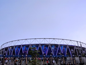 A large stadium with a circular design features multiple large banners displaying images of sports players in blue uniforms. The structure is modern, with a metal frame and triangular patterns. A crowd of people is gathered below, walking toward the entrance under a clear sky.
