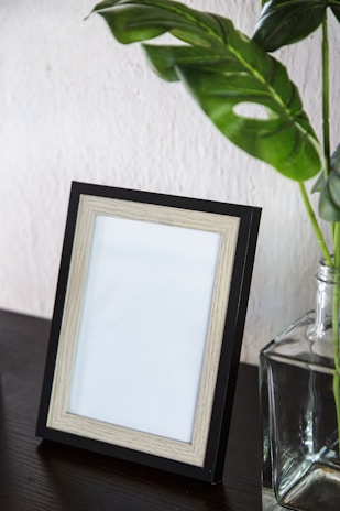 An empty picture frame with a black outer border and a lighter wood inner border, resting on a dark wooden surface. Next to the frame is a clear glass vase containing green leaves, adding a touch of nature to the setting. A textured white wall serves as the background.