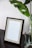 An empty picture frame with a black outer border and a lighter wood inner border, resting on a dark wooden surface. Next to the frame is a clear glass vase containing green leaves, adding a touch of nature to the setting. A textured white wall serves as the background.