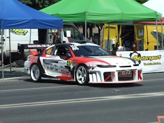 A race car is parked on a street next to tents and banners, with a design emphasizing red, white, and black. The car features various sponsor names and numbers, indicating it is set up for a motor sport event. There are tents and support vehicles in the background, suggesting a pit area at a racing event.