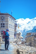 A person is standing with a backpack and trekking pole on a stone-paved path beside a rustic stone building. Snow-capped mountains and a clear blue sky are visible in the background. Nearby, traditional architecture and a white stupa with a golden pinnacle can be seen.