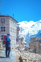 A person is standing with a backpack and trekking pole on a stone-paved path beside a rustic stone building. Snow-capped mountains and a clear blue sky are visible in the background. Nearby, traditional architecture and a white stupa with a golden pinnacle can be seen.