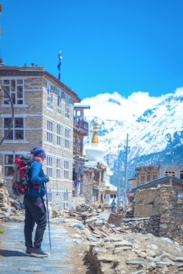A person is standing with a backpack and trekking pole on a stone-paved path beside a rustic stone building. Snow-capped mountains and a clear blue sky are visible in the background. Nearby, traditional architecture and a white stupa with a golden pinnacle can be seen.
