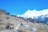 A mountainous village landscape with traditional stone houses positioned on terraces. The backdrop features snow-covered peaks under a bright blue sky. A few travelers walk along a path lined with dry shrubs, carrying supplies. Colorful prayer flags fly from poles scattered near the dwellings.