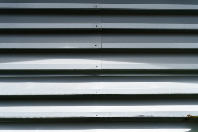 A metallic wall composed of horizontal ridges with a repetitive pattern. The surface has a smooth finish with visible shadows cast by the alternating ridges. Some signs of wear or minor discoloration can be observed on the lower edges.