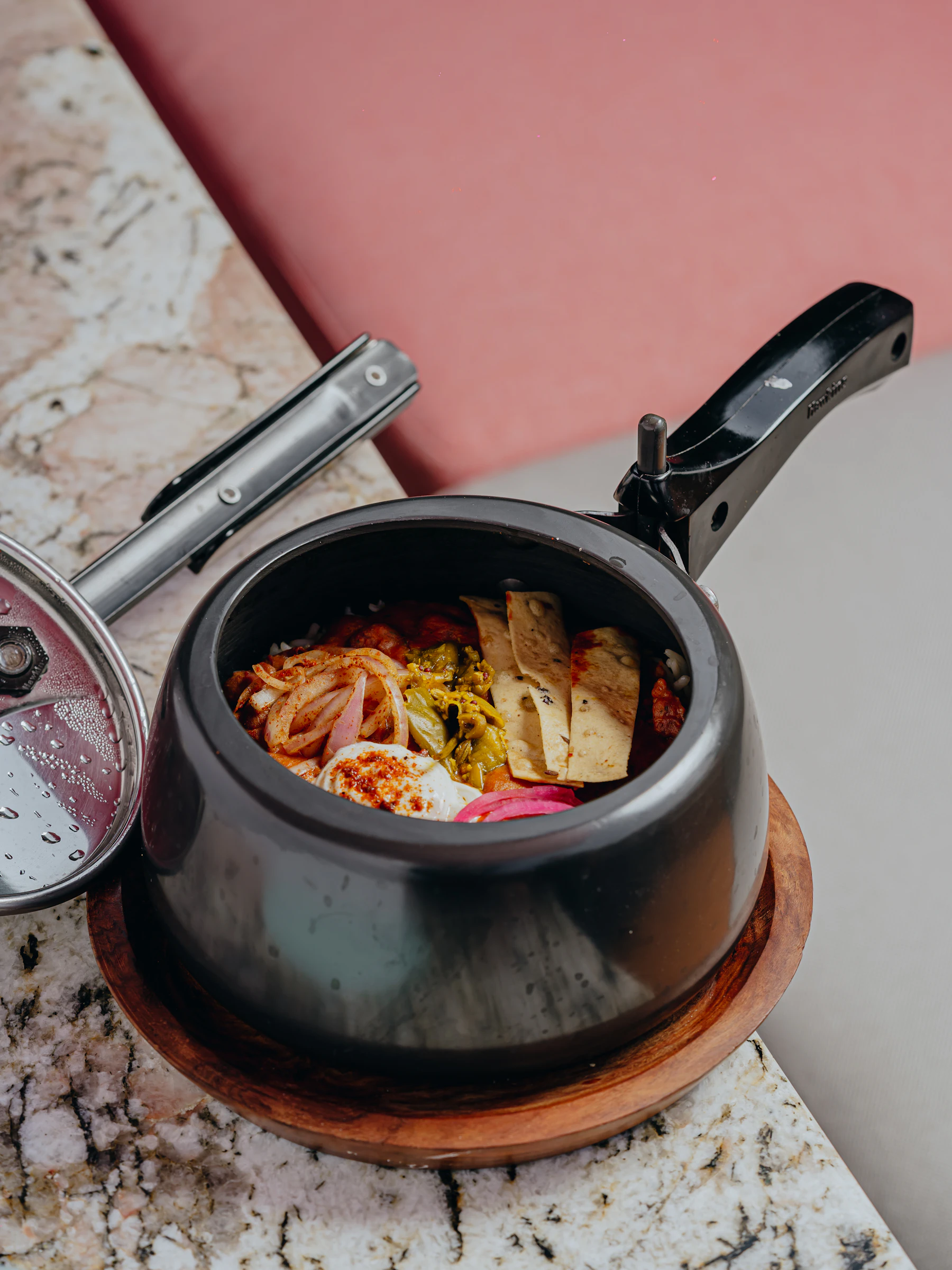 An open black stovetop pressure cooker full of food on a wooden stand, its locking lid lying beside it