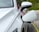 A white electric vehicle is parked and connected to a yellow charging cable. The car's side mirror and the charging port are clearly visible and the car is located in an outdoor setting with greenery in the background.