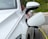 A white electric vehicle is parked and connected to a yellow charging cable. The car's side mirror and the charging port are clearly visible and the car is located in an outdoor setting with greenery in the background.
