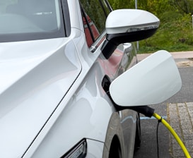 A white electric vehicle is parked and connected to a yellow charging cable. The car's side mirror and the charging port are clearly visible and the car is located in an outdoor setting with greenery in the background.