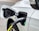 A close-up view of an electric vehicle being charged. The charging cable is securely attached to the port on the vehicle. The car is white, and the charging cable is predominantly black with a visible yellow section. There is a green indicator light on the charging port.