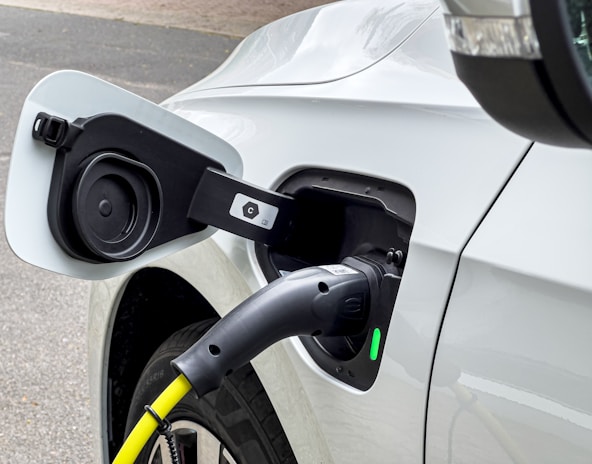 A close-up view of an electric vehicle being charged. The charging cable is securely attached to the port on the vehicle. The car is white, and the charging cable is predominantly black with a visible yellow section. There is a green indicator light on the charging port.