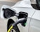 A close-up view of an electric vehicle being charged. The charging cable is securely attached to the port on the vehicle. The car is white, and the charging cable is predominantly black with a visible yellow section. There is a green indicator light on the charging port.