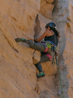 A person wearing climbing gear scales a rugged, beige rock face. The climber is equipped with a safety helmet and colorful climbing harness, carefully positioning their hands and feet in the crevices for support.