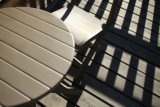 A wooden table and chair set on a deck with sunlight casting bold, dark shadows across the surface.