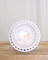 A close-up view of a modern LED light bulb placed on a wooden surface with a neutral backdrop. The bulb features a circular, grid-like design with a bright light at its center, surrounded by a white, perforated edge.