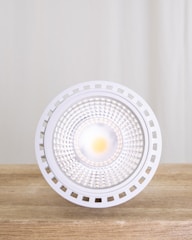 A close-up view of a modern LED light bulb placed on a wooden surface with a neutral backdrop. The bulb features a circular, grid-like design with a bright light at its center, surrounded by a white, perforated edge.
