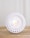 A close-up view of a modern LED light bulb placed on a wooden surface with a neutral backdrop. The bulb features a circular, grid-like design with a bright light at its center, surrounded by a white, perforated edge.