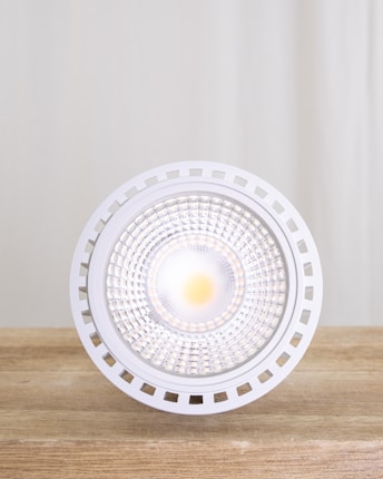 A close-up view of a modern LED light bulb placed on a wooden surface with a neutral backdrop. The bulb features a circular, grid-like design with a bright light at its center, surrounded by a white, perforated edge.