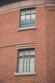 A section of a red brick wall with two large windows featuring metal frames and panes. The windows are set in a curved portion of the building, each with concrete sills. The architectural style highlights classic industrial design.