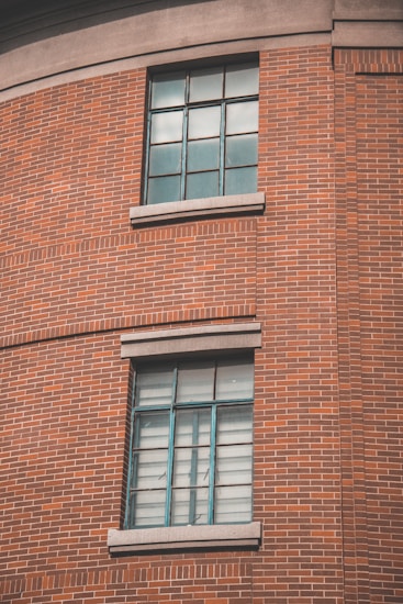 A section of a red brick wall with two large windows featuring metal frames and panes. The windows are set in a curved portion of the building, each with concrete sills. The architectural style highlights classic industrial design.
