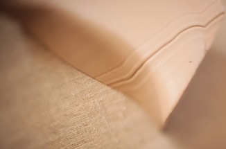 A close-up view of a textured fabric beside layers of paper with soft, beige tones. The fabric appears coarse and woven, while the paper has gentle curved edges.