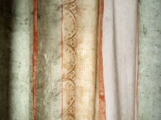 The image features a close-up of a textured fabric or curtain with varying vertical stripes. The central portion has a lace-like pattern with earth-toned colors such as beige and light brown. To the left and right, there are sections of greenish and off-white colors, with rusty orange accents adding contrast.
