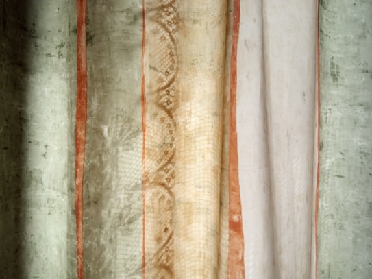 The image features a close-up of a textured fabric or curtain with varying vertical stripes. The central portion has a lace-like pattern with earth-toned colors such as beige and light brown. To the left and right, there are sections of greenish and off-white colors, with rusty orange accents adding contrast.