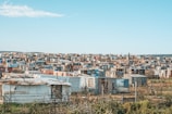 Numerous small, densely packed, makeshift houses and shanties spread out across a broad, semi-arid landscape under a clear blue sky. The structures are made from corrugated metal sheets, wood, and other scrap materials. The area appears to be a shantytown or informal settlement.
