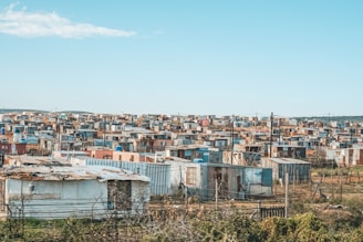 Numerous small, densely packed, makeshift houses and shanties spread out across a broad, semi-arid landscape under a clear blue sky. The structures are made from corrugated metal sheets, wood, and other scrap materials. The area appears to be a shantytown or informal settlement.