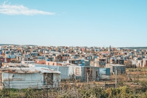 Numerous small, densely packed, makeshift houses and shanties spread out across a broad, semi-arid landscape under a clear blue sky. The structures are made from corrugated metal sheets, wood, and other scrap materials. The area appears to be a shantytown or informal settlement.