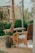 An outdoor seating area features wooden furniture, including a bench and a chair. A potted plant with orange flowers is placed near the wooden post supporting the structure. The background shows lush green bushes and partially visible wooden structures.