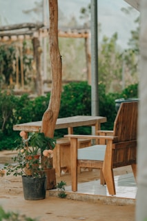 An outdoor seating area features wooden furniture, including a bench and a chair. A potted plant with orange flowers is placed near the wooden post supporting the structure. The background shows lush green bushes and partially visible wooden structures.