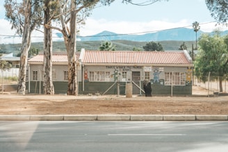 A small building with a red roof and light grey walls, surrounded by a fence. The front of the building features colorful murals of cartoon characters. There are several large trees in the foreground, and mountains appear in the background.