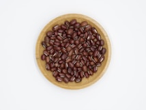 A wooden bowl filled with red beans is centered on a white background. The beans are small and have a shiny surface.