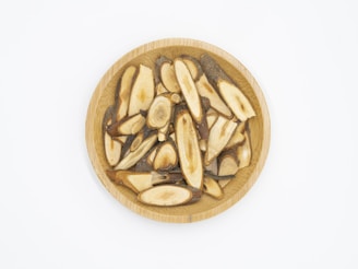A wooden bowl filled with sliced pieces of dried roots or bark, with smooth, beige interiors and dark edges. The bowl is centered against a plain white background, highlighting the natural textures and colors of the contents.