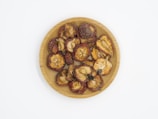 A wooden plate filled with dried fruit pieces, some with reddish edges and seeds visible in the center. The pieces have a wrinkled texture and varying shades of brown and red.