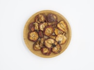 A wooden plate filled with dried fruit pieces, some with reddish edges and seeds visible in the center. The pieces have a wrinkled texture and varying shades of brown and red.