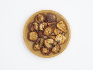 A wooden plate filled with dried fruit pieces, some with reddish edges and seeds visible in the center. The pieces have a wrinkled texture and varying shades of brown and red.