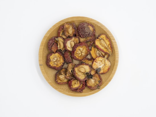 A wooden plate filled with dried fruit pieces, some with reddish edges and seeds visible in the center. The pieces have a wrinkled texture and varying shades of brown and red.
