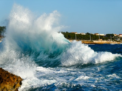 Large ocean wave crashing against rocky shore with foamy white spray under a clear blue sky. In the background, a coastline lined with trees and a row of houses.